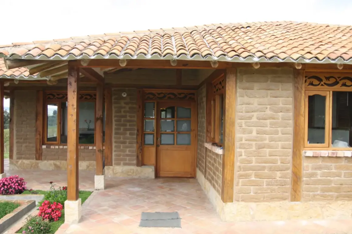 Fachada de casa con paredes de barro cocido, techo de tejas y detalles en madera. Flores rosadas y rojas adornan la entrada.