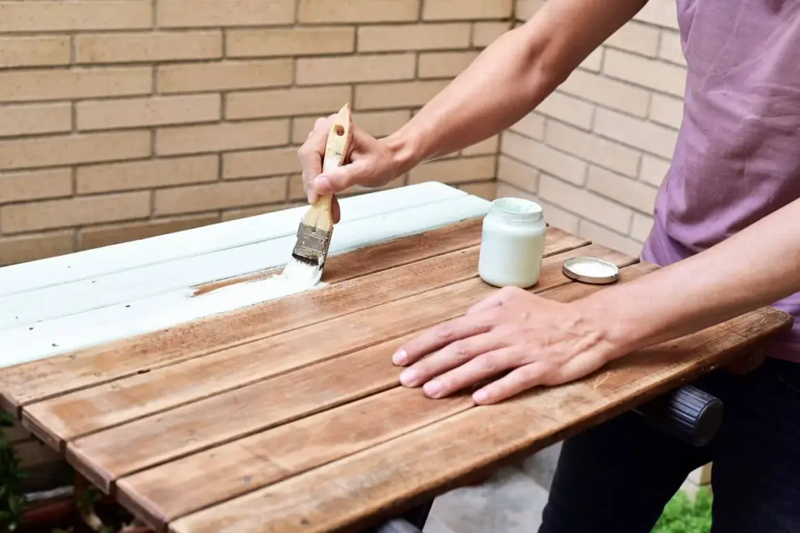 Persona aplica pintura blanca a una mesa de madera ya pintada. El proceso de como pintar madera ya pintada es sencillo con esta técnica.