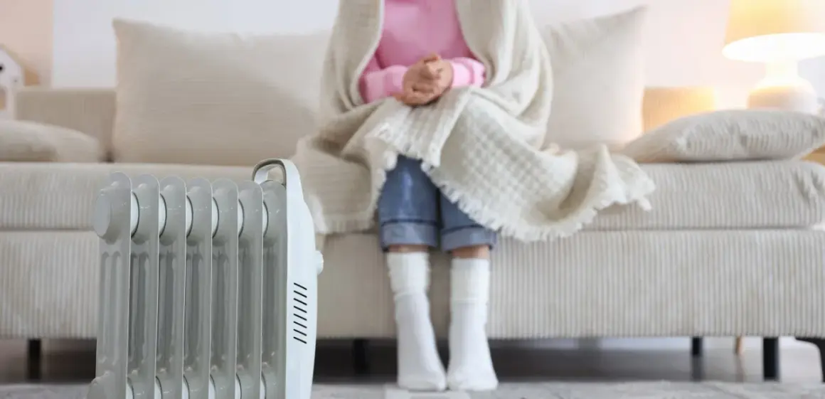Persona abrigada con manta y calcetines sentada en sofá, junto a un radiador eléctrico, solución de calefacción sin instalación.