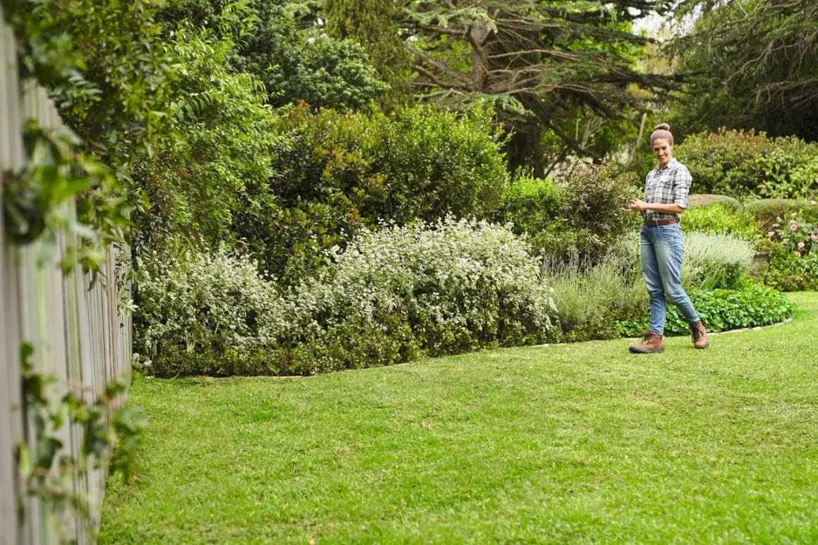 Mujer sonríe en un jardín exuberante, con un seto de arbustos blancos y verdes, y un gran chopo al fondo.
