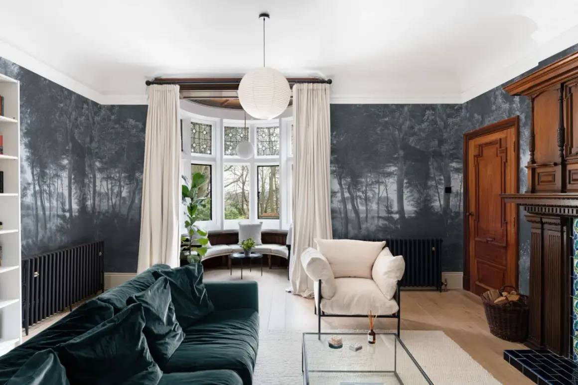 Un salón con papel pintado de bosque oscuro, un sofá verde, sillón beige y chimenea de madera. Ideas para cómo empapelar una pared con estilo.