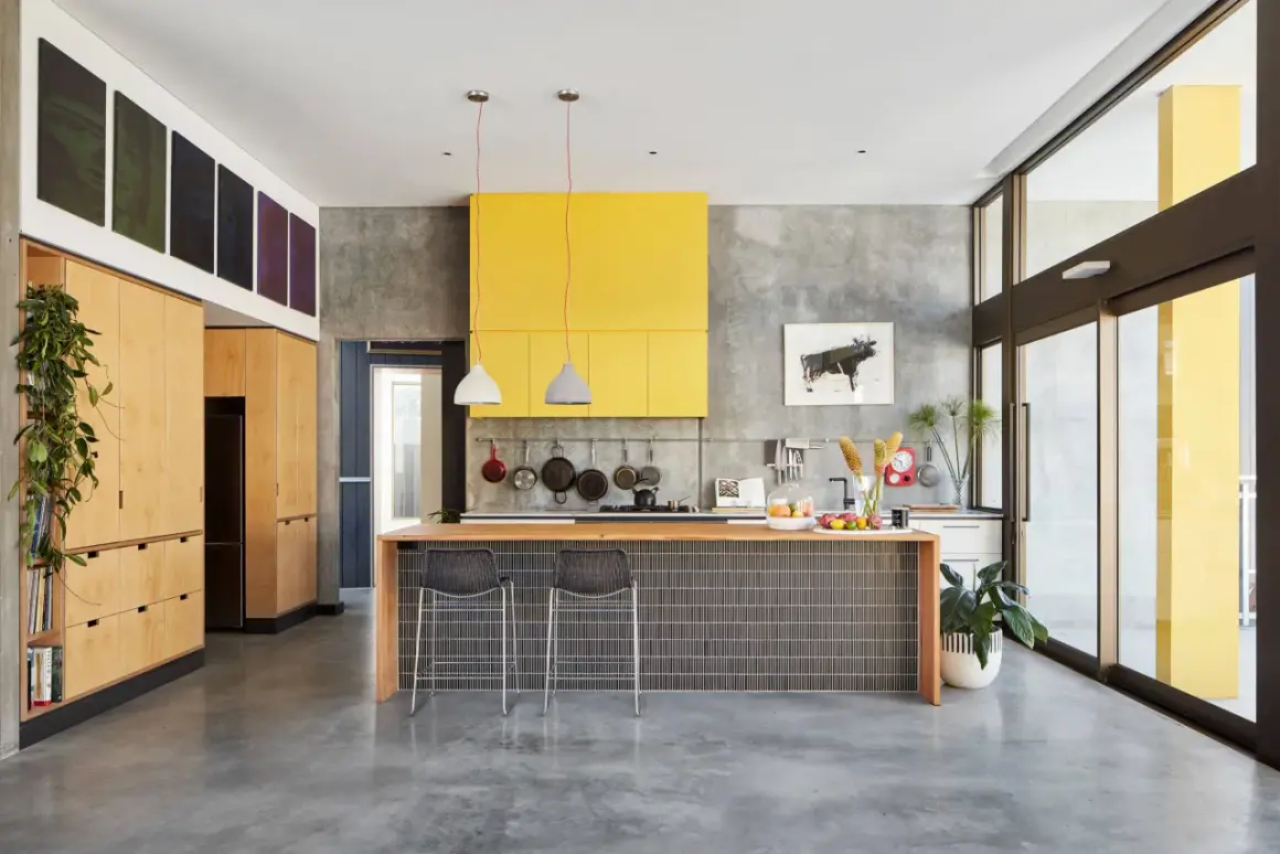 Cocina moderna con suelo de cemento pulido, isla central con azulejos, campana amarilla y armarios de madera.