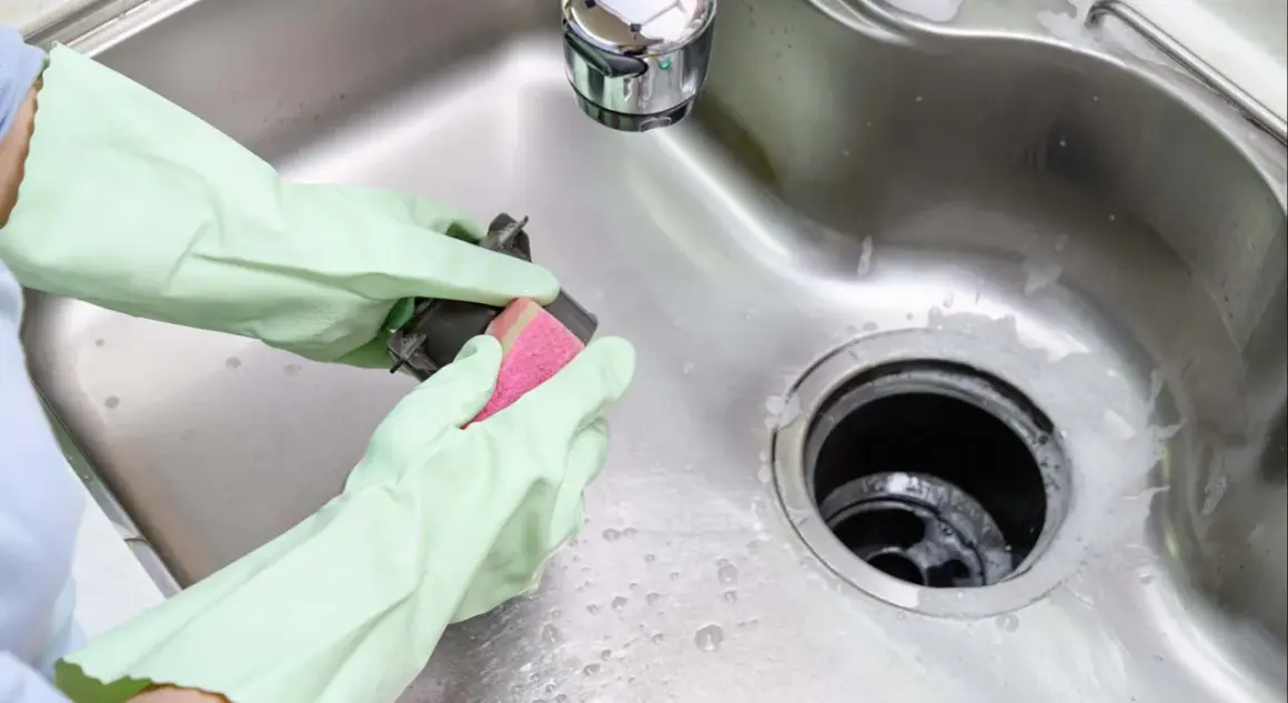 Manos enguantadas limpian un triturador de basura con una esponja rosa, un desatascador casero improvisado para evitar atascos en el fregadero.