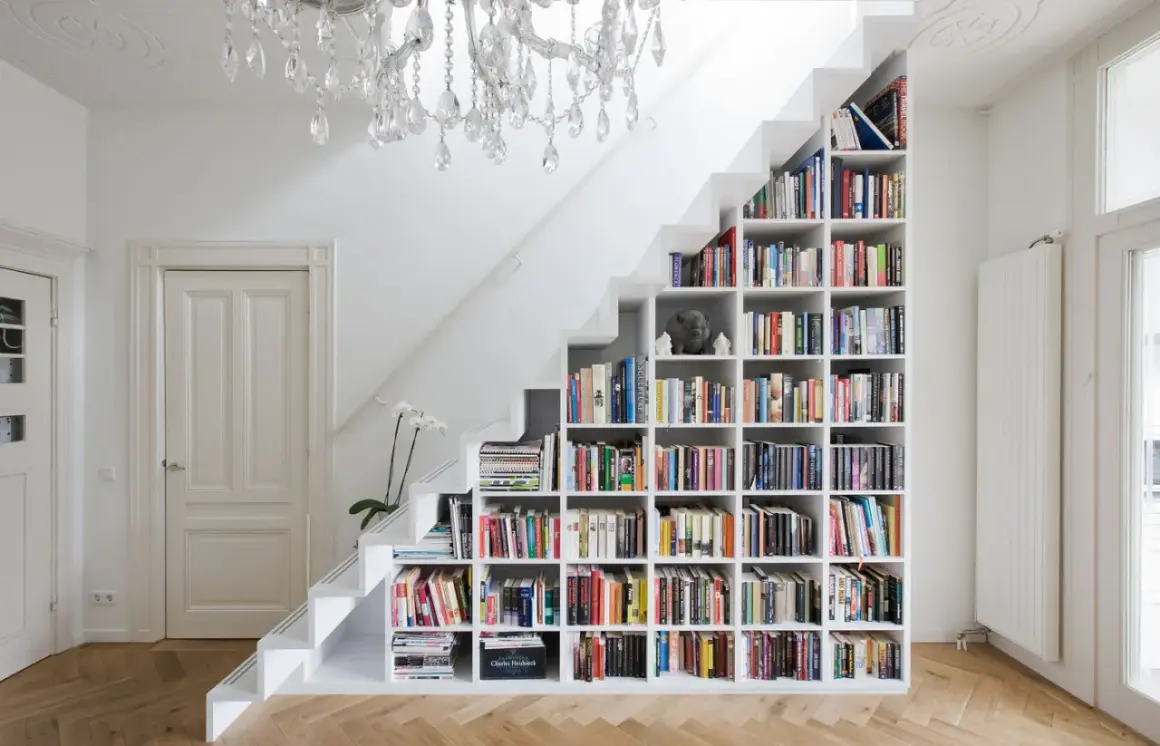 Estantería blanca de pladur para cerrar hueco bajo escalera, llena de libros y con una lámpara de araña.