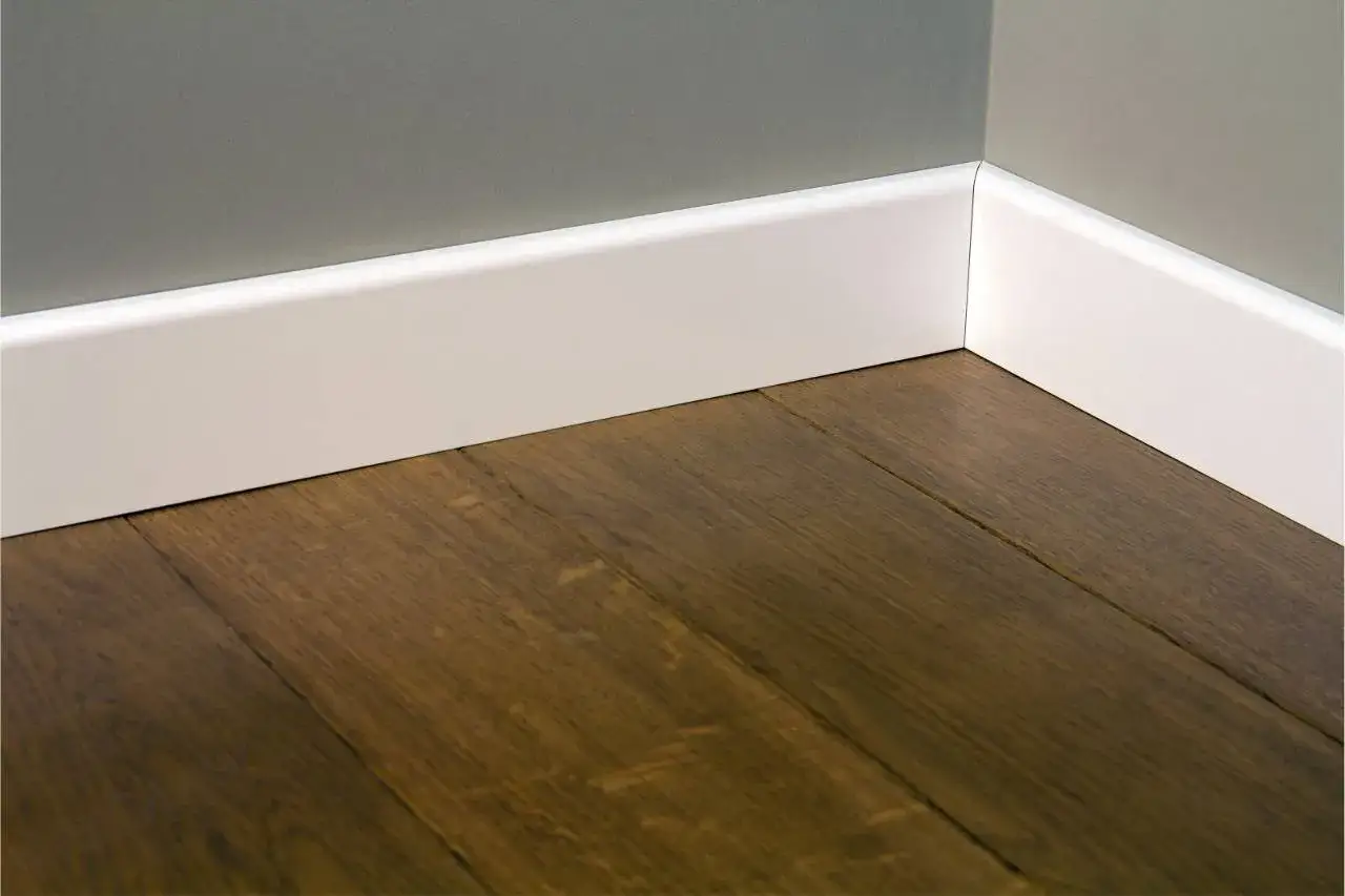 Esquina de una habitaci&oacute;n con suelo de madera oscura y rodapi&eacute;s blancos modernos. Hay varios tipos de rodapi&eacute;s, y este es un ejemplo minimalista.