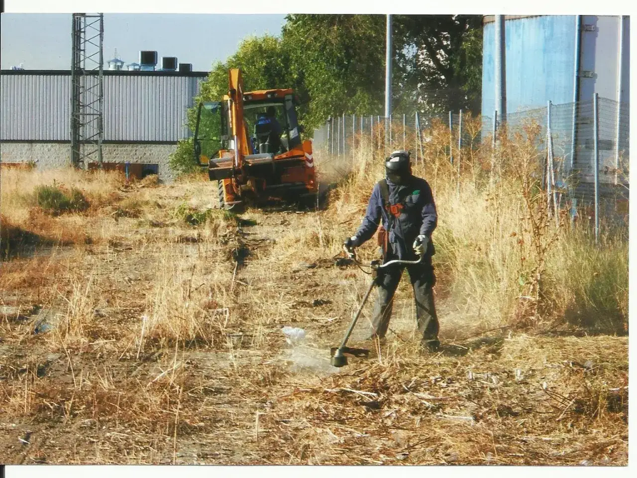 Hombre con desbrozadora realiza desbroce de terreno. Una retroexcavadora naranja está estacionada cerca.