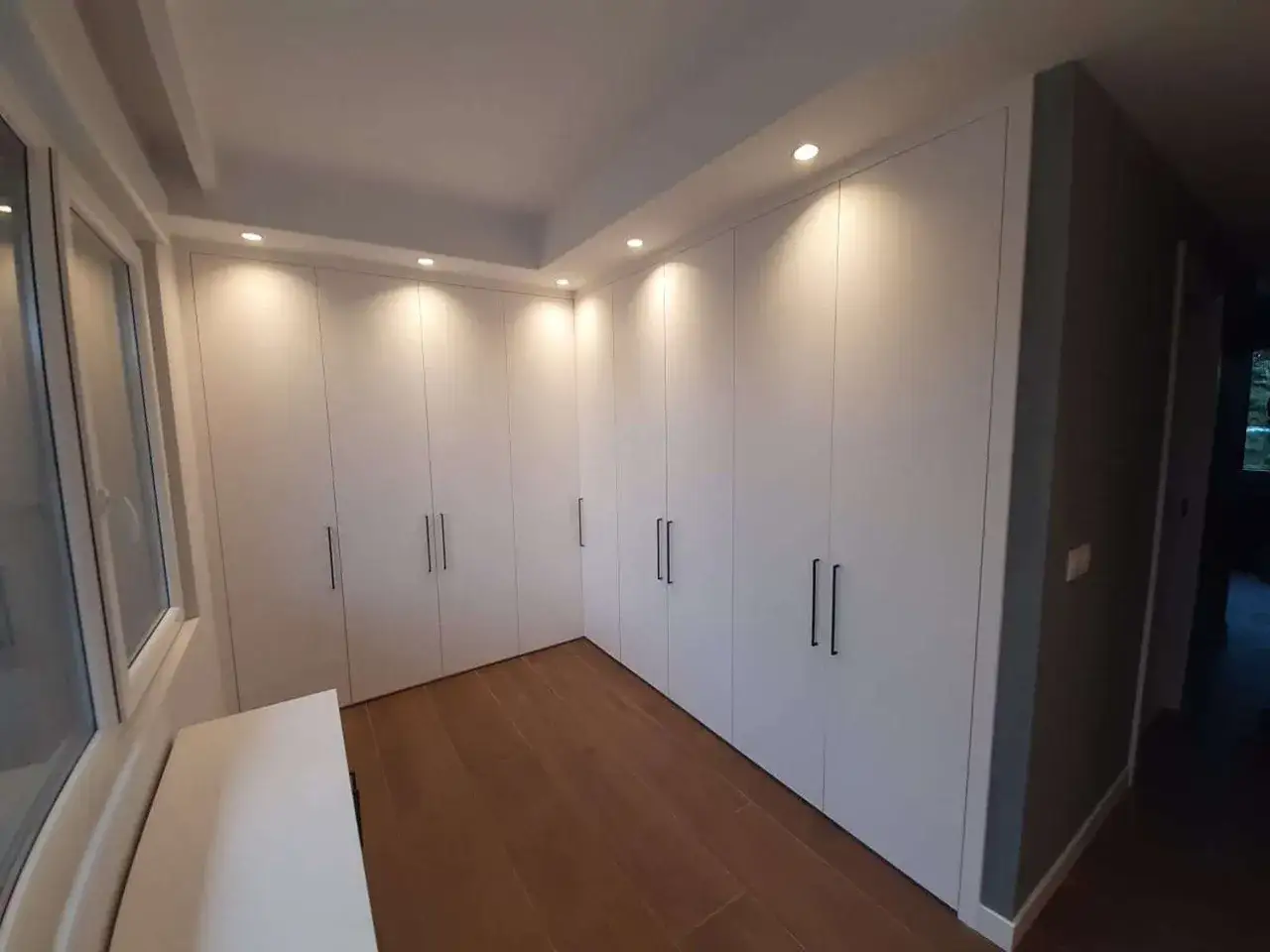 Amplio vestidor cerrado con puertas blancas y tiradores negros. Suelos de madera y luz empotrada en el techo.
