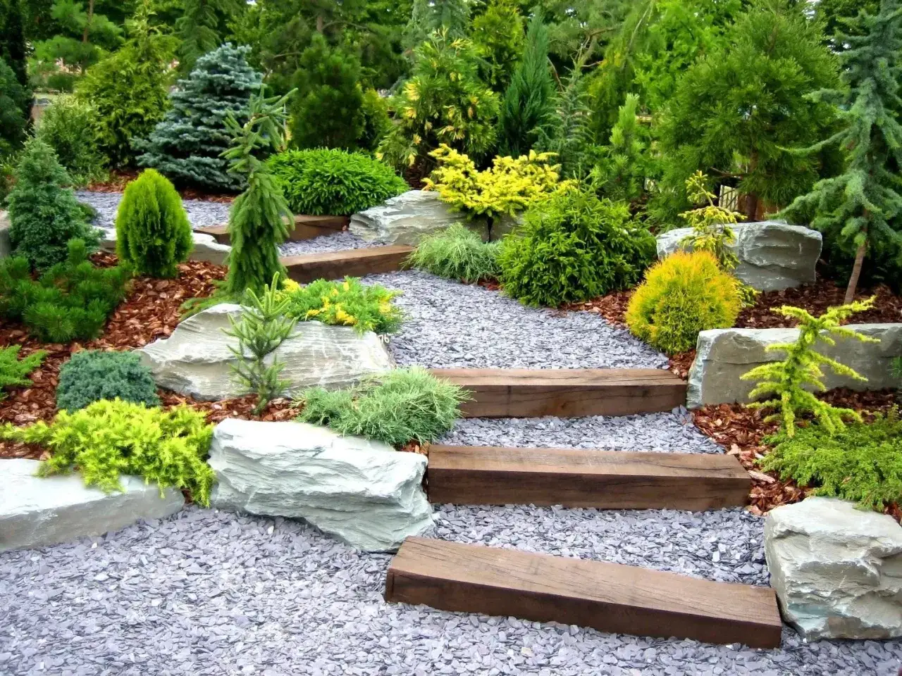 Un sendero serpenteante entre jardines con piedras y escalones de madera, rodeado de coníferas y arbustos verdes y amarillos.
