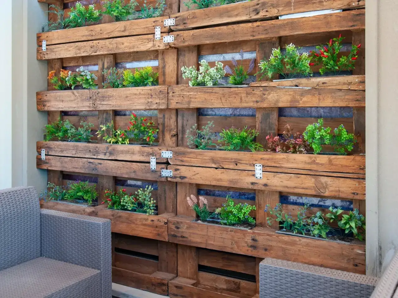 Un jard&iacute;n vertical con palets lleno de plantas verdes y flores de colores alegra un rinc&oacute;n exterior, junto a un sill&oacute;n gris.