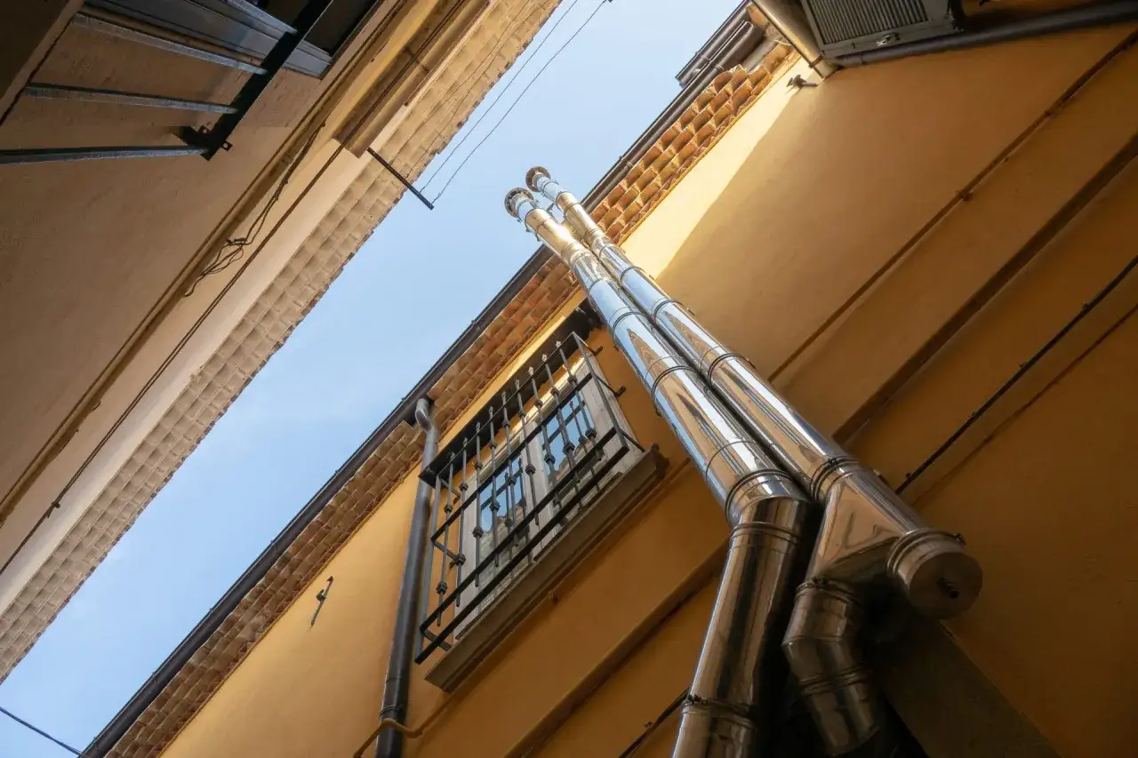 Instalaci&oacute;n estufa pellets: dos tubos de acero inoxidable ascienden por una fachada amarilla, saliendo de una ventana con rejas.