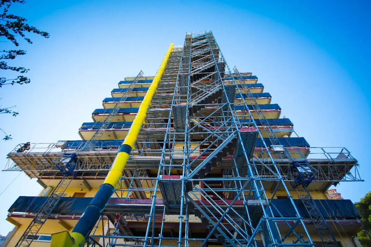 Edificio en construcci&oacute;n con varios tipos de andamios met&aacute;licos y un tubo de desescombro amarillo.