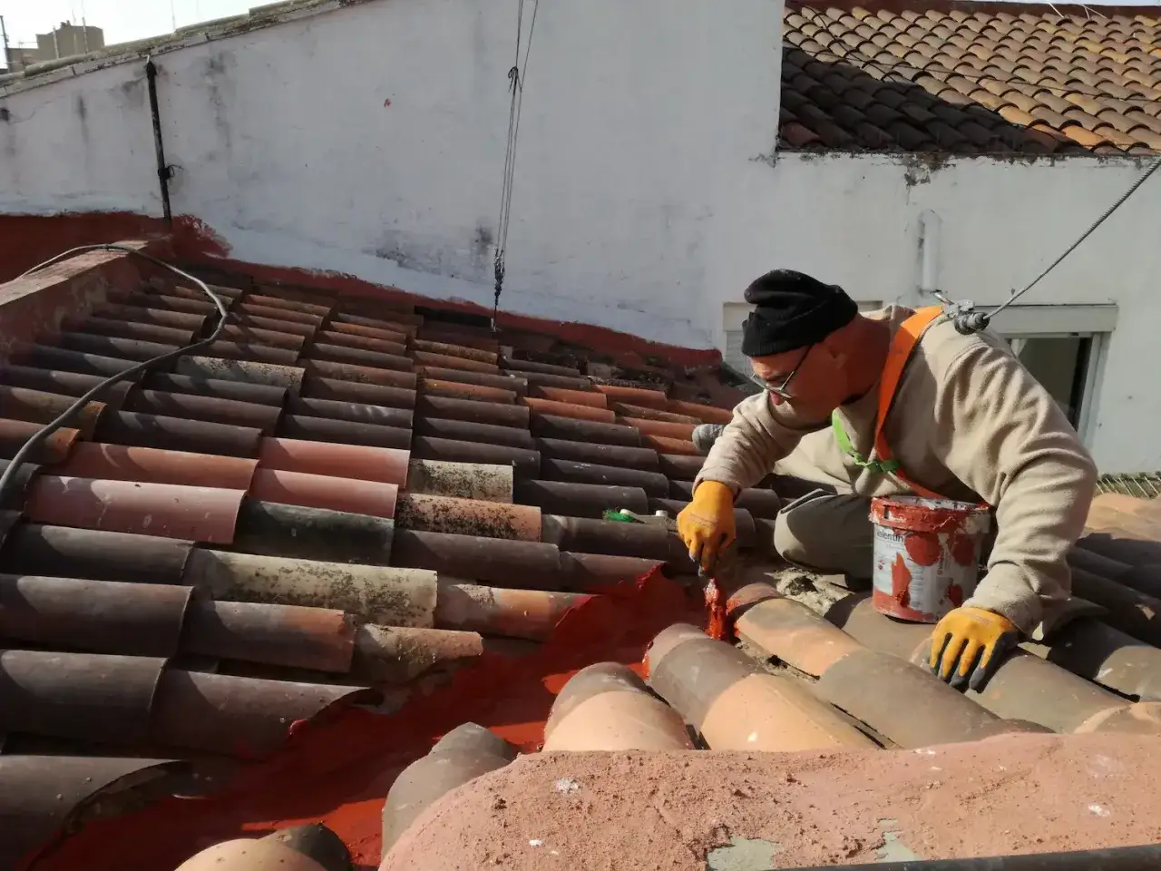 Hombre con guantes naranjas y gorro negro aplica pintura roja para reparar tejado.