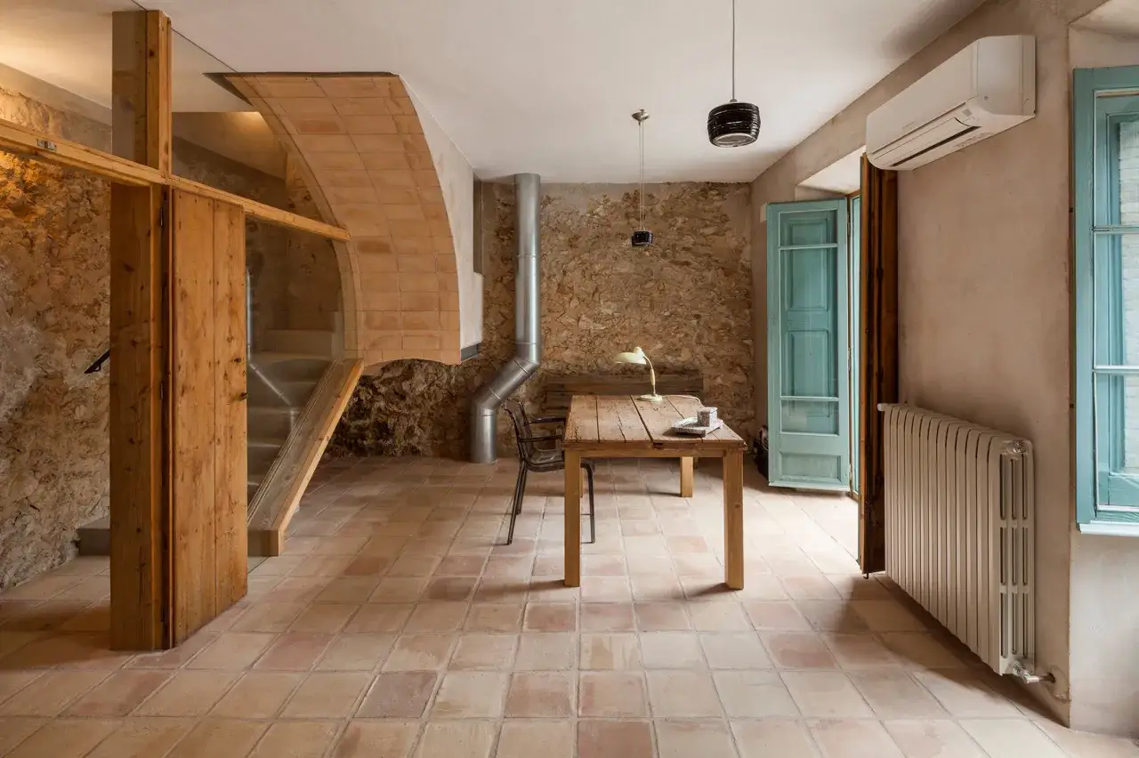 Suelo de barro cocido en un espacio r&uacute;stico con paredes de piedra, escalera de madera y una mesa de trabajo.