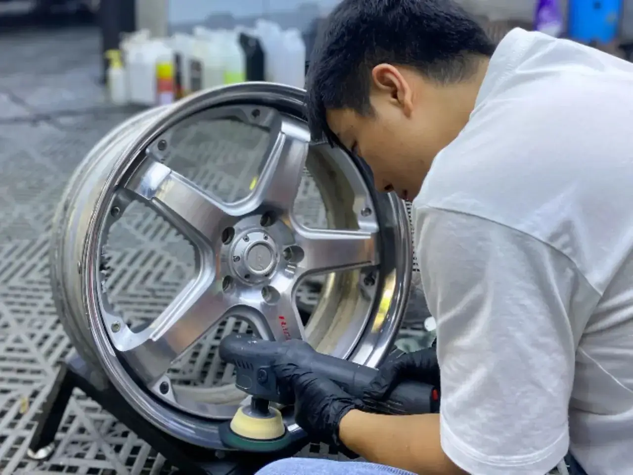 Hombre con guantes negros usa una pulidora para pulir aluminio de una llanta de coche, dejándola reluciente.