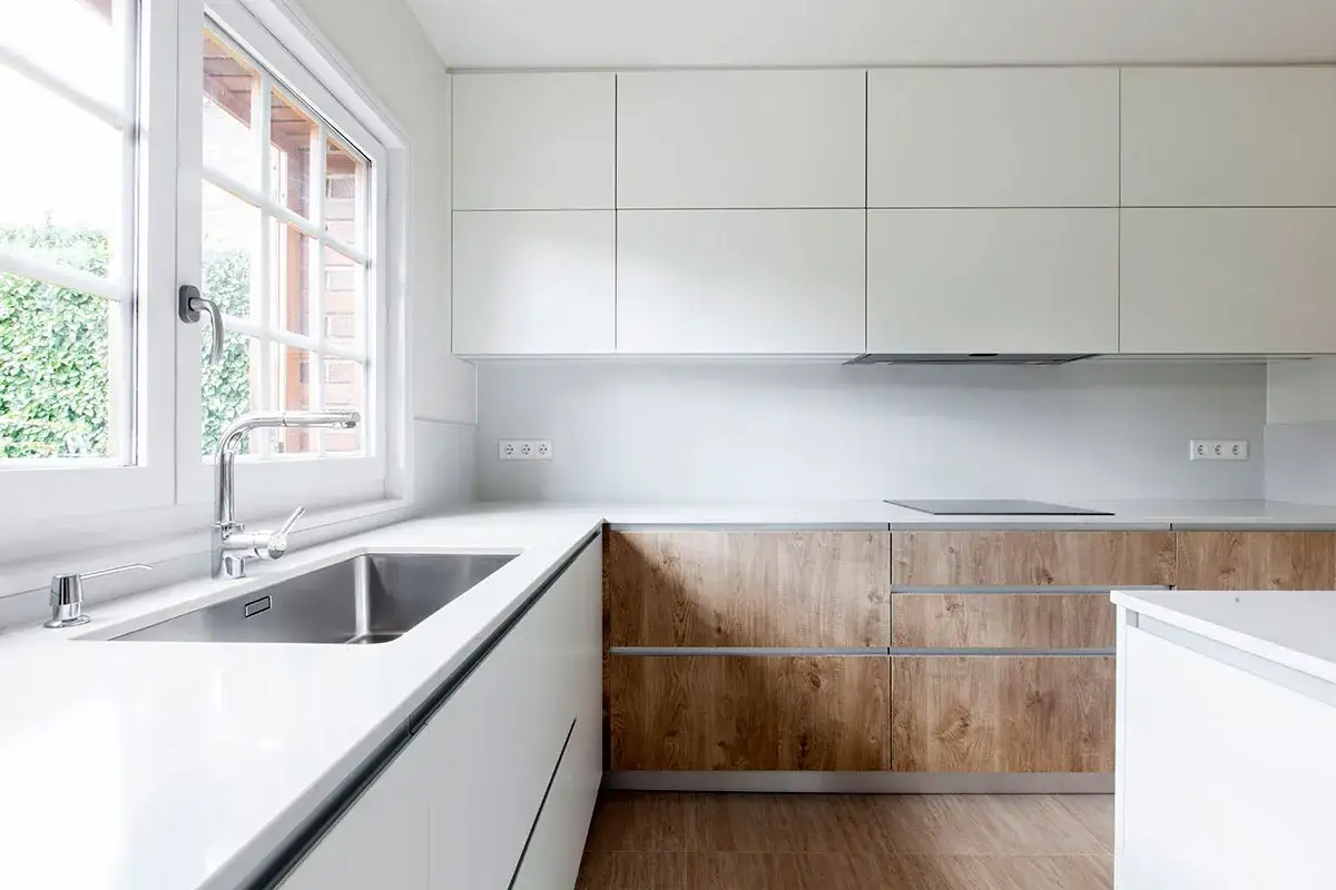 Cocina moderna con muebles de melamina o lacado blanco y detalles en madera. Encimera blanca, fregadero de acero inoxidable y ventana con vistas al jardín.