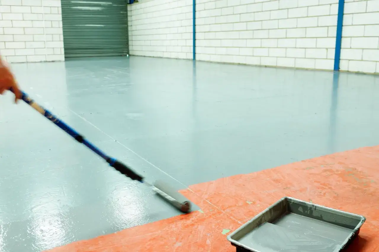 Persona aplica pintura gris a un suelo industrial con un rodillo. La bandeja de pintura est&aacute; sobre una l&aacute;mina naranja.