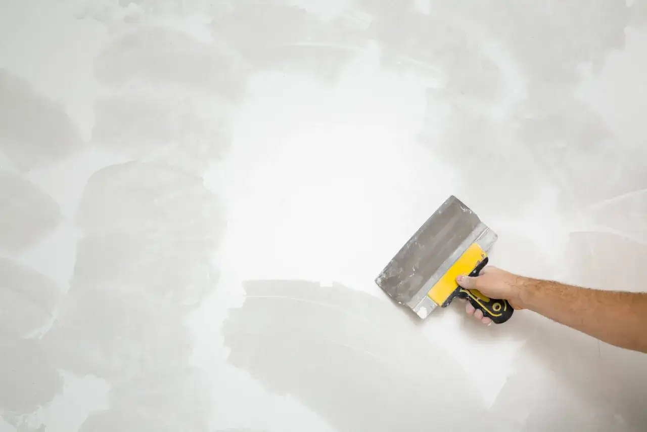 Mano con esp&aacute;tula amarilla y gris alisando una pared con gotel&eacute;, mostrando c&oacute;mo quitar el gotel&eacute; sin esfuerzo.