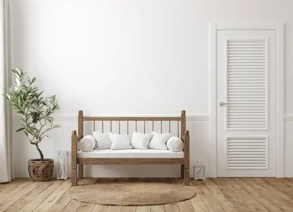 Un banco de madera rústico con cojines blancos, junto a una planta y una puerta blanca. Ideal para aprender como hacer un banco de madera.