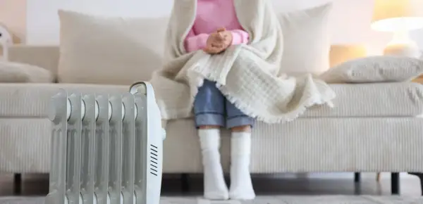 Persona abrigada con manta y calcetines sentada en sofá, junto a un radiador eléctrico, solución de calefacción sin instalación.