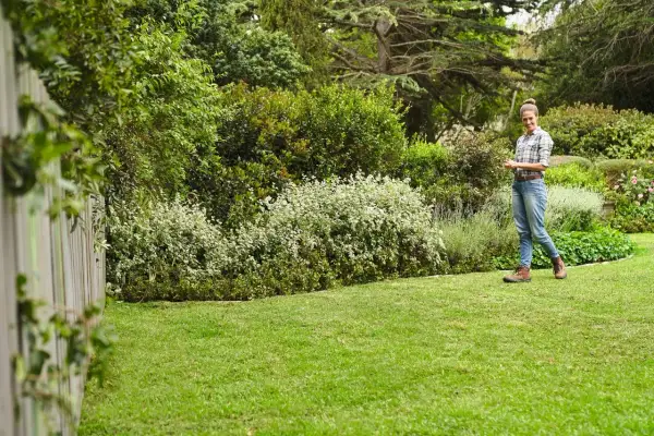 Mujer sonríe en un jardín exuberante, con un seto de arbustos blancos y verdes, y un gran chopo al fondo.