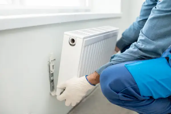 Man con guantes blancos ajusta un radiador blanco en la pared. Se está llevando a cabo la tarea de cambiar radiador de sitio.