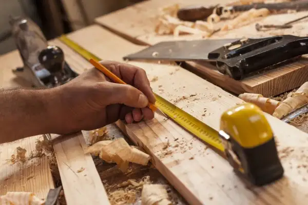 Mano marcando madera con lápiz y cinta métrica, lista para empezar a hacer una estantería de madera. Herramientas de carpintería esparcidas.